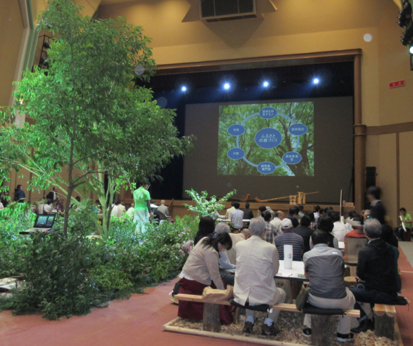 会場には大きな木が飾られており、スクリーンに映し出されている「ふるさとの森づくり」について座ってみている人々の写真