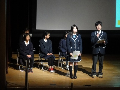 舞台上に制服を着た生徒が座り、その前に2名の男女生徒が紙を持ち話をしている写真