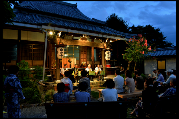 お寺の前で4人の男性が横並びで座りサックスを演奏している写真