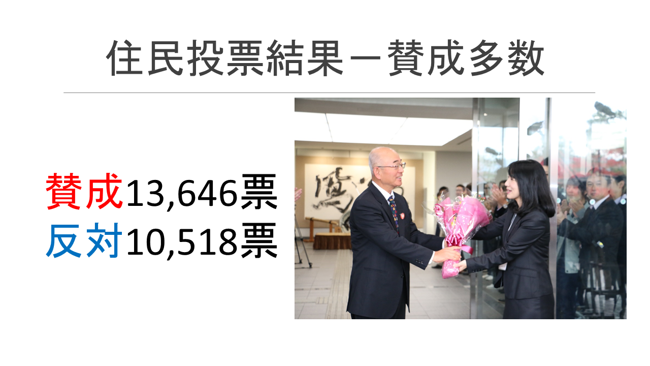 住民投票結果―賛成多数（賛成13,646票、反対10,518票）と書かれたスライドに秘書から酒井市長に向かって花束を贈っている写真